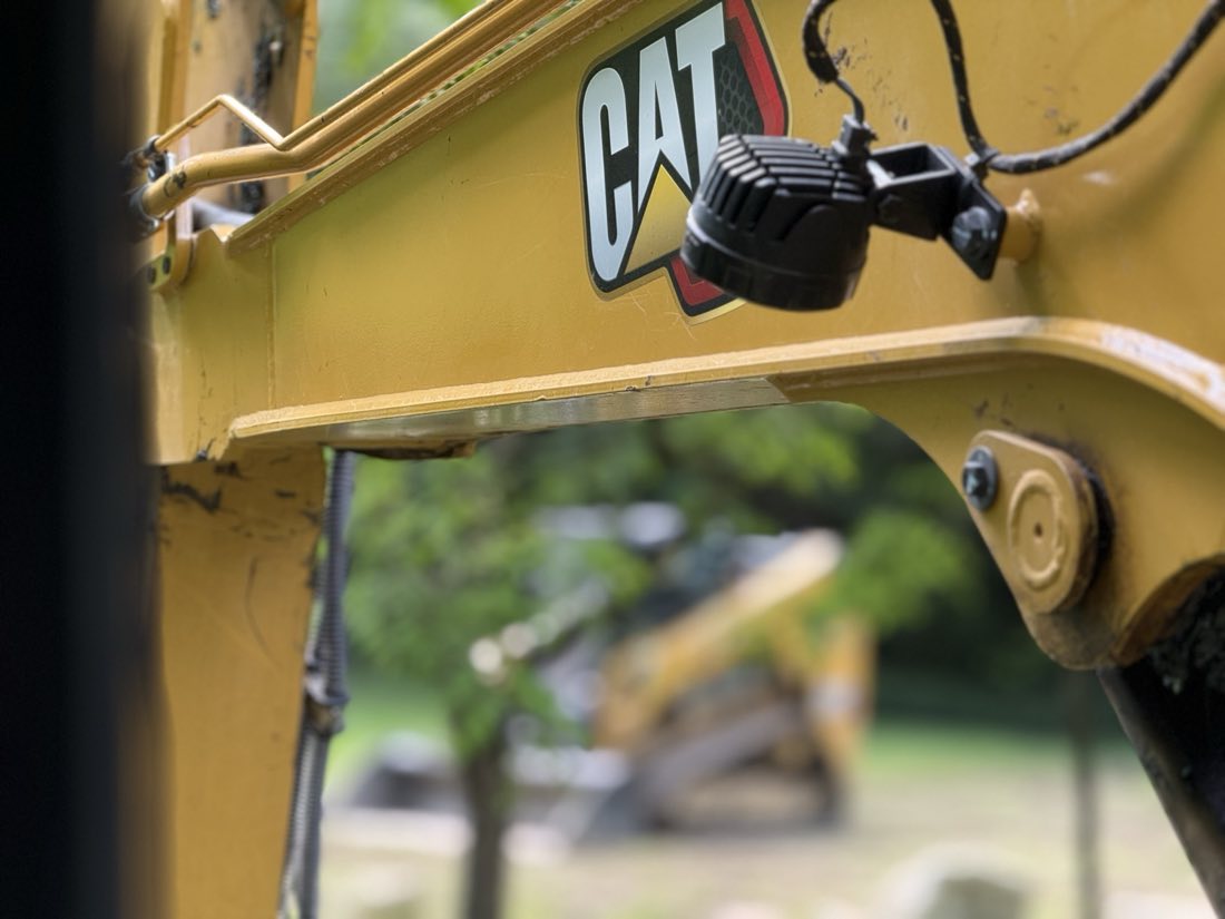 Close detail of a CAT skid steer's boom arm and work light, with a second machine visible out of focus in the background.