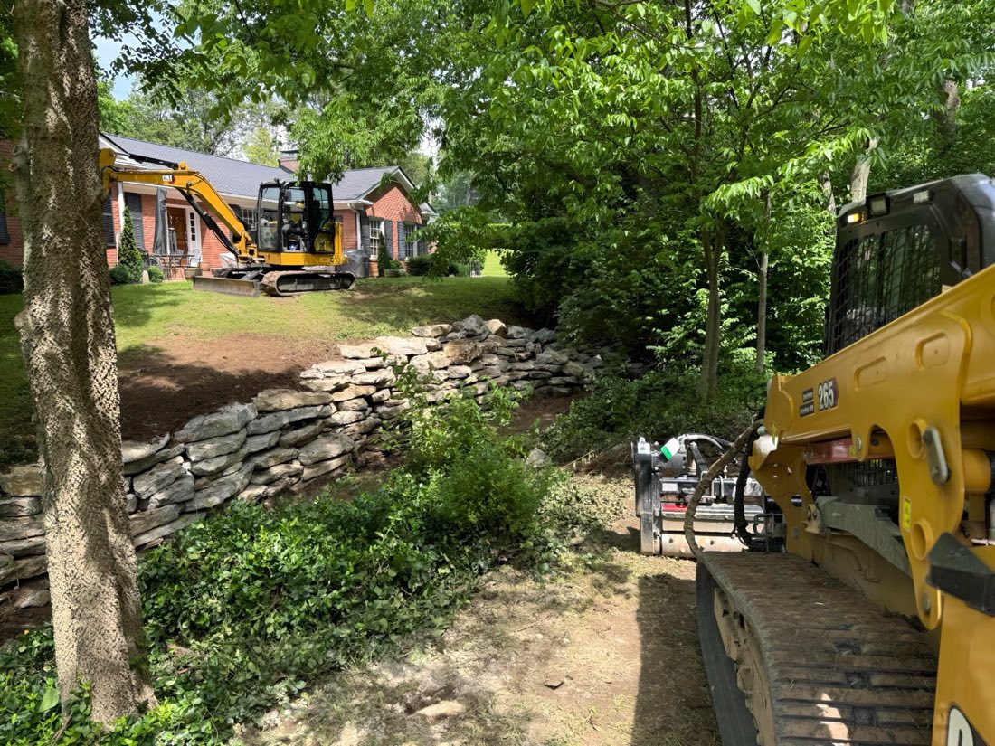 A CAT mini excavator and a CAT compact track loader staged on a residential property, brick house and a dry-stone retaining wall behind them.