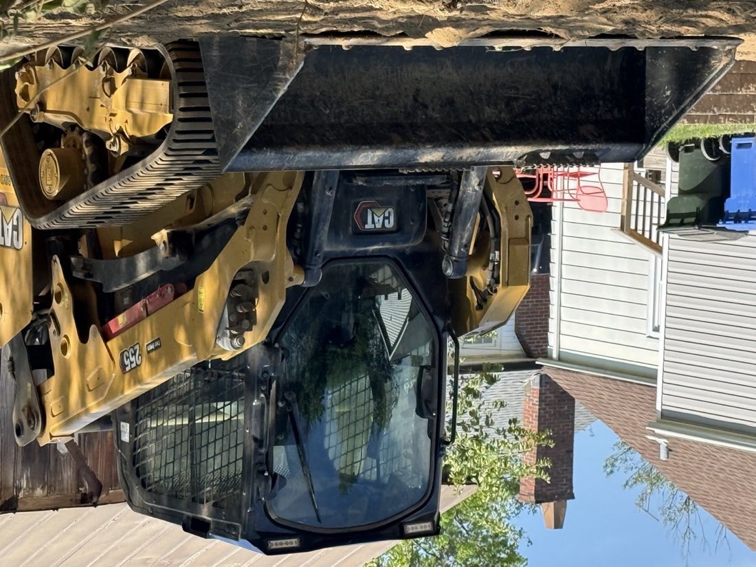 A CAT compact track loader on an active residential jobsite, houses and a fenced yard visible behind it.