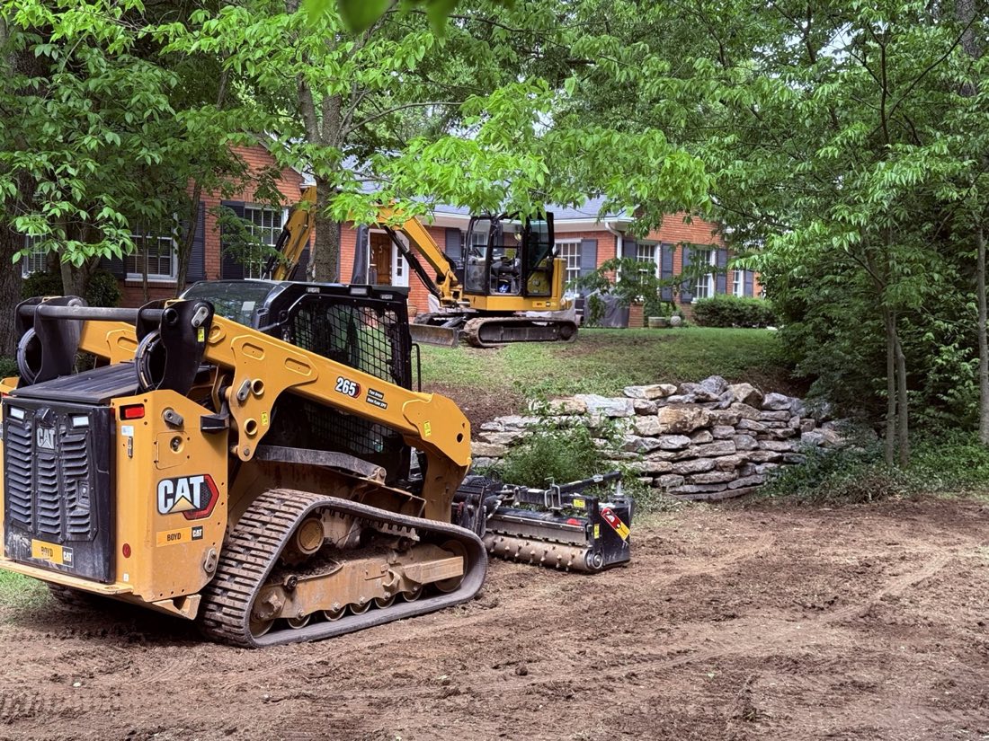 Compact track loader and mini excavator working a wooded residential property, with a completed dry-stone retaining wall along a drainage swale.